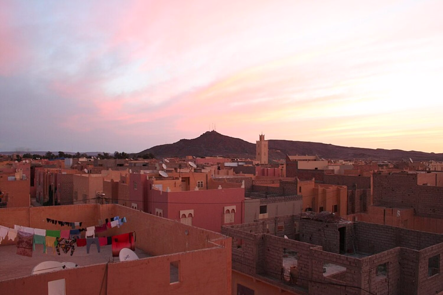 Vista del atardecer sobre la ciudad desértica de Erfoud en el sureste de Marruecos, con casas de tejados planos y una colina árida al fondo.
