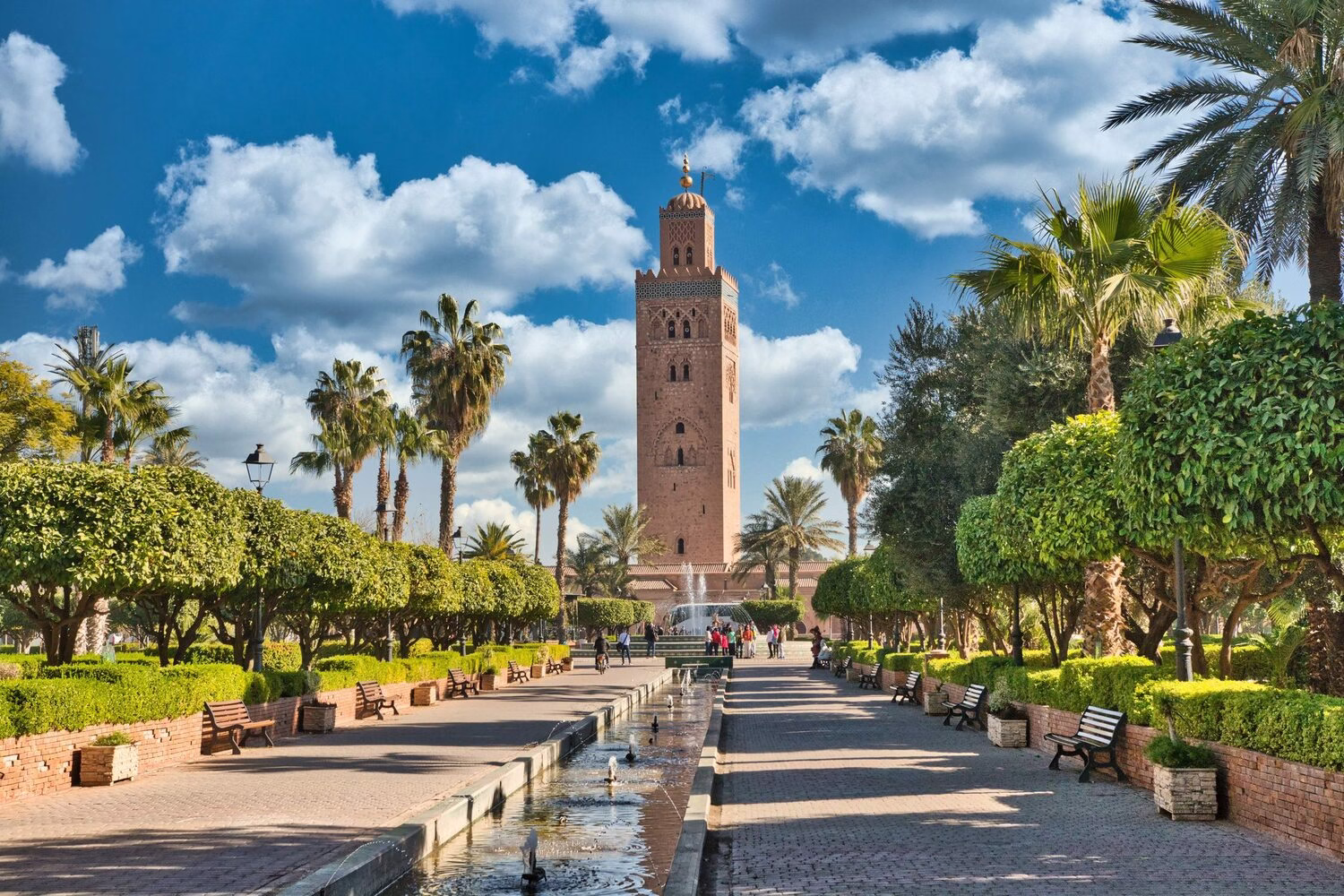 Mezquita Kutubía en Marrakech con su icónico minarete, jardines bordeados de palmeras, fuentes y un cielo azul claro en el corazón de la ciudad